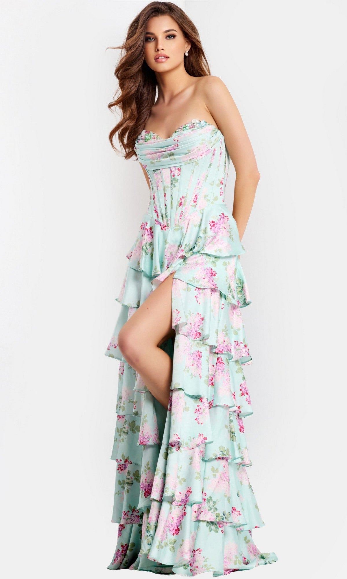 A woman wears the Jovani 47579 long prom dress in print, featuring a strapless corset bodice, tiered skirt, pink floral design, and thigh-high slit, posing against a plain light background.